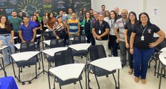 #PraTodosVerem: a fotografia aparecem pessoas em pé, em uma sala de aula, com diversas carteiras...