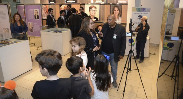 #PraTodosVerem: Na fotografia aparece crianças em uma fila, ao fundo uma mesa com uma urna eletr...