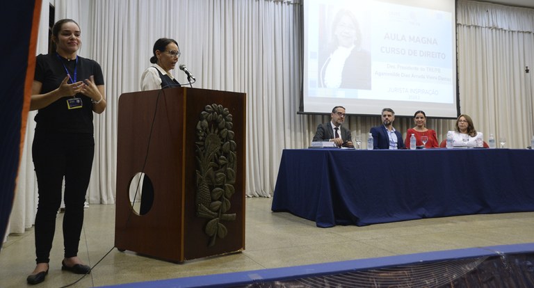 Painel retangular, no primeiro plano, uma mulher fala do púlpito; em segundo plano, pessoas sent...