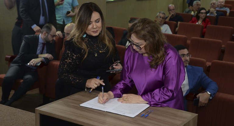 Duas mulheres de pé, uma delas assinando um documento, em evento da Justiça Eleitoral.