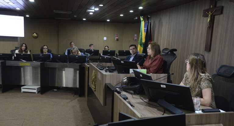 Painel retangular com pessoas sentadas à mesa de julgamento, em reunião.