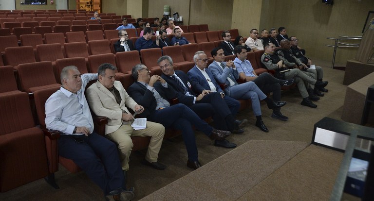 Painel retangular com pessoas sentadas em cadeiras de auditório, em reunião.