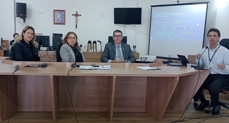 Painel retangular com quatro pessoas sentadas à mesa de reunião, dois homens e duas mulheres; no...