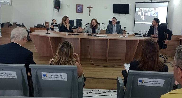 Painel retangular com quatro pessoas sentadas à mesa de reunião, dois homens e duas mulheres; em...