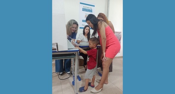 #PraTodosVerem: Na fotografia aparecem três mulheres, auxiliando uma criança a votar, em uma seç...